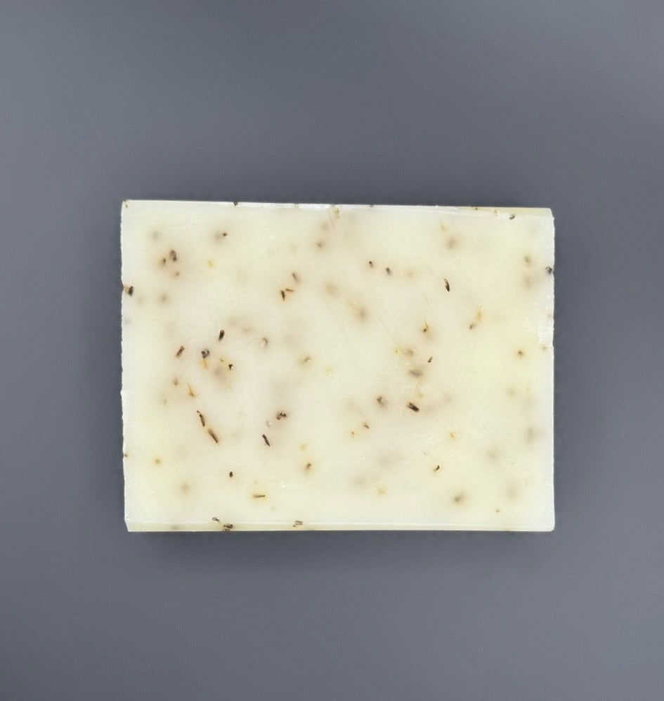 A rectangular bar of handmade soap with a creamy off-white base and scattered small brown botanical inclusions, shown on a solid dark gray background.
