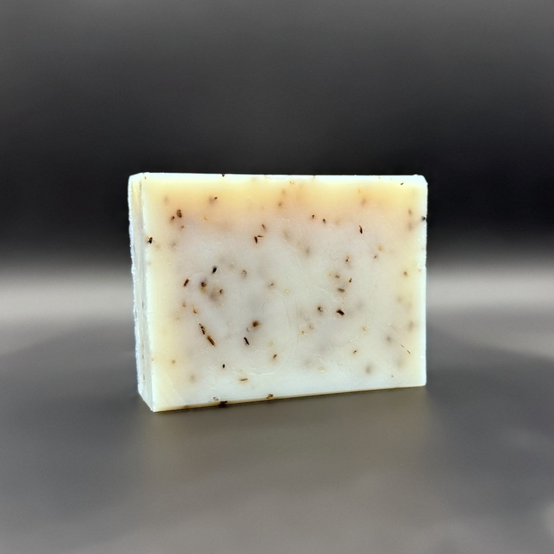 A rectangular bar of handmade soap with a creamy off-white base and scattered dark brown botanical inclusions, standing upright on a dark gray reflective surface.