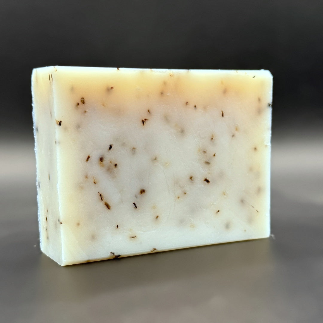 A rectangular bar of handmade soap with a creamy off-white base and scattered dark brown botanical inclusions, photographed on a dark gray gradient background.