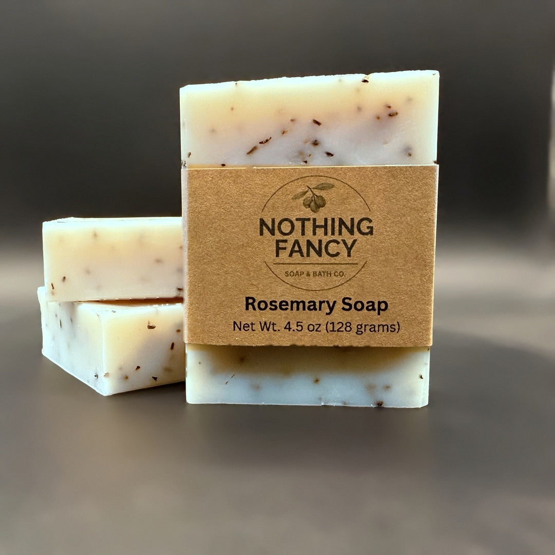 Three bars of Nothing Fancy Rosemary Soap stacked on a dark gray surface, with the front bar wrapped in a kraft paper label.