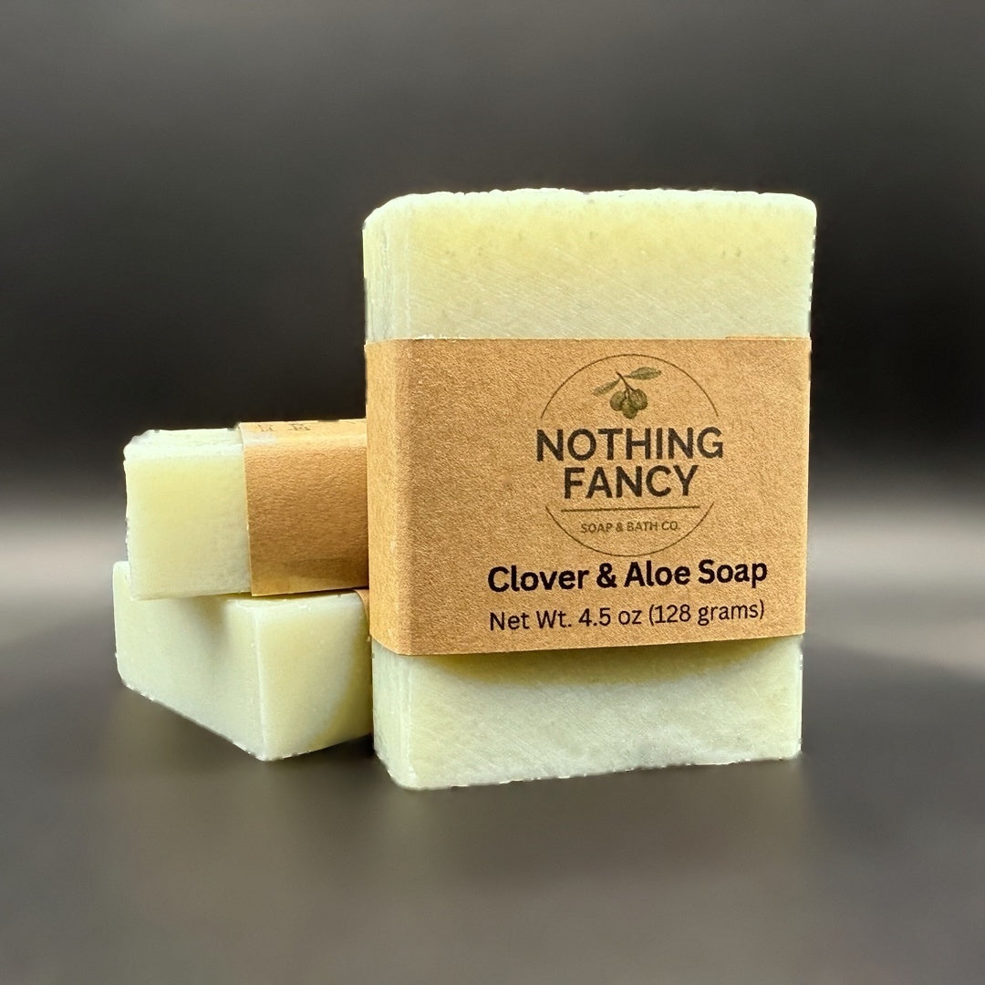 A bar of soap wrapped in a kraft paper band labeled 'NOTHING FANCY Clover & Aloe Soap' stands in front of two unwrapped pale green soap bars on a dark gray background.
